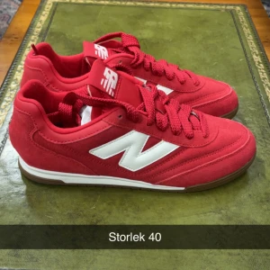 Röda New Balance sneakers strl 40 - Snygga röda sneakers från New Balance med vit logga på sidan och matchande röda snören. Skorna har en låg profil och platt sula, tillverkade i mocka och syntet. Perfekta för dig som gillar sportig stil och vill sticka ut med färg.