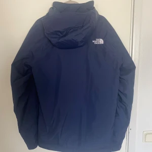 The North Face vinterjacka - Snygg marinblå jacka från The North Face med huva och dragkedja framtill. Jackan har en stilren design med logga på bröstet.. Perfekt för dig som vill ha en clean look och skydd mot regn samt håller värmen!