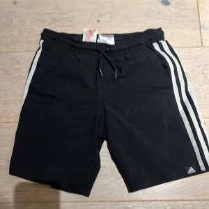 Svarta Adidas shorts med ränder - Svarta shorts från Adidas med klassiska vita ränder längs sidorna. De har elastisk midja med snörning och är gjorda i lätt polyester, perfekt för sport och träning. Snygg och enkel design med Adidas-logga på benet.