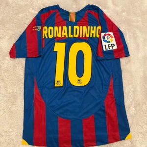 FC Barcelona Ronaldinho tröja Nike - Klassisk FC Barcelona fotbollströja med Ronaldinho och nummer 10 på ryggen. Tröjan är blå och röd med gula detaljer, randigt mönster och officiella klubbmärken samt LFP-patch på ärmen. Tillverkad i lätt och ventilerande polyester.