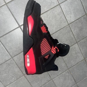 Nike Air Jordan 4 svart/röd sneakers - Säljer ett par Nike Air Jordan 4 sneakers i svart och rött med klassisk Jumpman-logga på hälen. Skorna har meshdetaljer, snörning och en synlig Air-enhet i sulan. Ovandelen är i mocka och syntet, med coola röda accenter och svart yttersula.