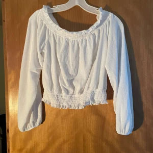 Offshoulder blus med volang och smock - Säljer en söt vit blus med lång ärm och volangdetaljer vid halsringningen. Blusen har smockad midja och är tillverkad i ett lätt, strukturerat tyg som ger en luftig känsla. Perfekt för dig som gillar romantisk stil och vill ha något fräscht i garderoben.