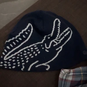 Mörkblå Lacoste mössa med krokodil - Snygg mörkblå mössa från Lacoste med stor vit krokodil på sidan. Perfekt för dig som gillar streetstyle och vill sticka ut med ett coolt mönster. Mjuk och stickad för extra komfort under kyliga dagar.
