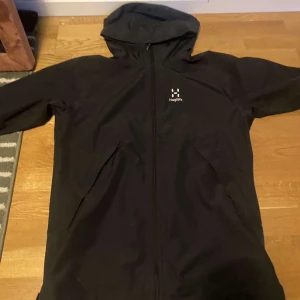 Svart regnjacka från Haglöfs med huva - Svart regnjacka från Haglöfs i storlek S. Jackan har en stilren design med hel dragkedja framtill, justerbar huva och två sidofickor. Materialet är syntet och jackan är långärmad med normal passform.
