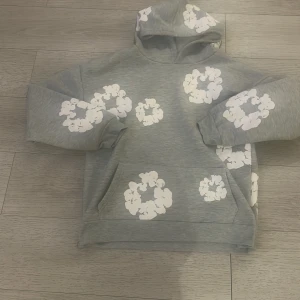 Grå hoodie med blommor från Denim Tears - Grå hoodie från Denim Tears med vita blommiga tryck över hela tröjan. Tröjan har huva, känguruficka och långa ärmar. Mjuk insida och avslappnad passform, perfekt för en chill look.