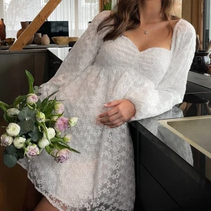 Vit kortklänning från NA-KD - Superfin vit kortklänning från NA-KD med puffiga långärmar och söt hjärtformad urringning. Klänningen har ett blommigt spetsmönster, perfekt för sommarens alla tillfällen som student, midsommar osv!