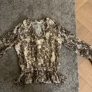 Leopard blus från Howtokissafrog  - Säljer en transparent leopard blus från Howtokissafrog i beige och bruna toner. Blusen har långa ärmar, snörning vid halsen och volangdetalj nedtill. Materialet är lätt och luftigt, perfekt för vår och sommar. Supersnygg till jeans eller kjol!