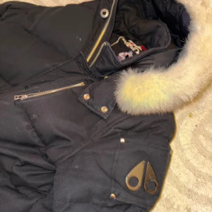 Svart dunjacka Moose Knuckles L=Large - Säljer en svart dunjacka från Moose Knuckles med pälskantad huva och flera fickor med dragkedja och knappar. Jackan har en klassisk logga på ärmen och quiltad design som håller värmen under kalla dagar. Perfekt för vintern!