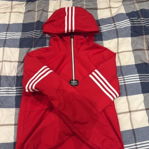 Röd Adidas vindjacka med huva - Säljer en röd Adidas vindjacka med klassiska vita ränder på ärmar och huva. Jackan har en half zip framtill, huva med dragsko och elastiska muddar. Materialet är lätt och syntetiskt, perfekt för blåsiga dagar. Adidas Originals-logga på bröstet.