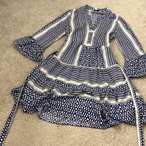 Blåvit mönstrad kortklänning Pigalle XS - Supersnygg kortklänning från Pigalle i blått och vitt med grafiskt mönster. Klänningen har volangärmar, v-ringning och markerad midja med knytband. Materialet känns lätt och luftigt, perfekt för varmare dagar. 