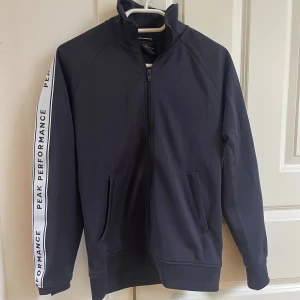 Mörkblå Peak Performance hoodie - Snygg mörkblå hoodie från Peak Performance med dragkedja och höga krage. Längs ärmarna finns vita band med logotypen tryckt i svart. Tröjan har två fickor framtill och är tillverkad i mjuk bomullsmix, perfekt för chill eller träning.