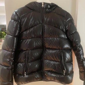 Svart pufferjacka från Colmar - Svart pufferjacka från Colmar med glansig finish och huva. Jackan har två sidofickor med dragkedja, quiltad design och Colmar-logga på ärmen. Perfekt för kalla dagar och har en riktigt snygg streetstil. Dragkedja framtill och långärmad modell.