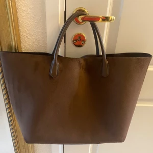 Brun shopper i mockaimitation från H&M - Det finns kortare handtag och ett par längre handtag att fälla upp för att kunna hänga den på axeln.   Använd endast ett par gånger så den är i mycket bra skick.   Mått: höjd utan handtag: 30 cm Bredd: 48 cm Bottenplatta: 34 x 13 cm  Mörkbrun Mocka imitation HM Axelväska Handväska Totebag Tote bag Shopperbag Shoppingväska kaffebrun 