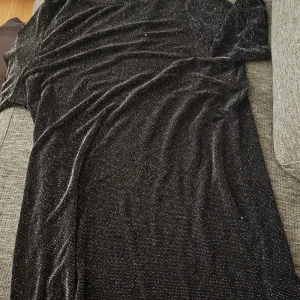 Svart glittrig långklänning Monki - Svart långklänning från Monki med glittriga silvertrådar som ger en snygg shimmer-effekt. Klänningen har en rak och enkel passform och är perfekt för dig som vill sticka ut med en stilren look. Materialet är mjukt och följsamt, vilket gör den bekväm att bära.