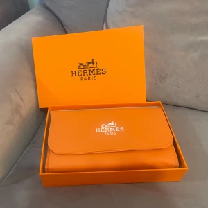 Väska för (sminkborstar) - Snygg orange kuvertväska med klassisk logga på framsidan. Väskan är rektangulär och har plats för sminkborstar eller andra småsaker. Tillverkad i skinn med slät yta och praktisk knappstängning. Perfekt för dig som gillar lyxiga accessoarer. Med borstarna!!!!!