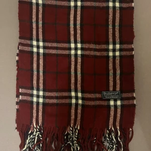 Rutig vinröd halsduk från Burberry - Snygg halsduk från Burberry i klassiskt rutigt mönster med vinröd bas och inslag av vitt och svart. Gjord i 100% lammull, med fransar i ändarna som ger en cool touch. Perfekt accessoar för att lyfta din outfit under kyliga dagar. halsduken skickad från Norge och moms kan tilkomma. betalning måste ske via paypal eller liknande