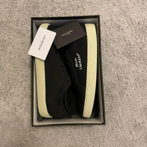 Svarta Saint Laurent sneakers - Snygga svarta sneakers från Saint Laurent med vit sula och logotypen broderad på sidan. Skorna har klassisk snörning och en stilren, låg profil. Perfekta för dig som gillar minimalistisk design och exklusiva märken.