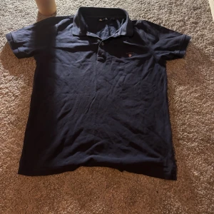 Mörkblå pikétröja från GANT Storlek 146/152 12år - Snygg mörkblå pikétröja från GANT med klassisk krage och två knappar framtill. Tröjan har broderad GANT-logga med sköld på bröstet och är tillverkad i mjuk bomull. Perfekt för en chill och stilren look.