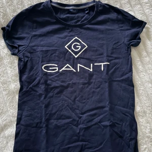 Mörkblå GANT t-shirt med logga - Snygg mörkblå t-shirt från GANT i mjuk bomull. Klassisk rund hals och vit GANT-logga tryckt framtill. Perfekt passform med korta ärmar och stilren design som funkar till jeans eller shorts.
