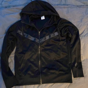 Svart hoodie från Nike med dragkedja - Svart hoodie från Nike med hel dragkedja framtill och huva med snörning. Snyggt mönstrad banddetalj över bröstet med Nike-logga. Tröjan har två fickor fram och långa ärmar med Nike-logga på vänster ärm. Perfekt för chill eller träning.