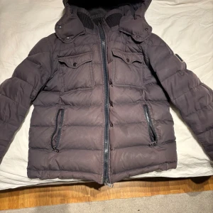 Mörkgrå Moncler pufferjacka med huva - Säljer en äkta mörkgrå pufferjacka från Moncler med huva och ribbade muddar. Jackan har två bröstfickor, samt två sidofickor med dragkedja. Insidan är fodrad med glansigt material och har en innerficka med dragkedja. Perfekt för kalla dagar.