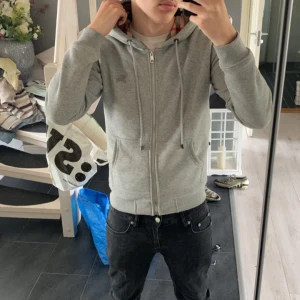 Grå Burberry hoodie med dragkedja - Säljer en grå hoodie från Burberry med klassiskt rutigt foder i huvan. Tröjan har dragkedja, snörning vid huvan och två fickor framtill. Perfekt för dig som gillar stilrena detaljer och vill ha något som sticker ut med det ikoniska Burberry-mönstret.