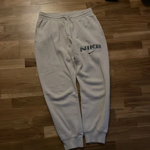 Vita mjukisbyxor från Nike med broderad logga - Säljer ett par vita mjukisbyxor från Nike i storlek M. Byxorna har broderad Nike-logga på benet, elastisk midja med snörning och muddar vid bensluten. Bra skick inte använda mer än ett fåtal gånger. 