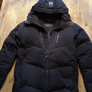 Svart pufferjacka från sail racing (2800kr ny) - Svart pufferjacka från Sail racing med huva och två bröstfickor med dragkedja. Storleken är 160 men på bilderna är jag 175 cm lång och den passar bra! Jackan har några små tecken på användning men bara använd ett fåtal gånger