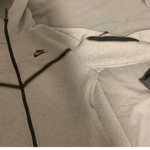 Grå Nike hoodie med svarta detaljer - Snygg ljusgrå hoodie från Nike med svarta dragkedjor och detaljer. Tröjan har huva, hel dragkedja framtill och en extra ficka med dragkedja på ärmen. Perfekt för chill eller träning, klassisk Nike-logga på bröstet.