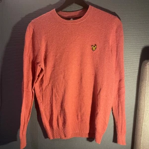 Rosa/röd tröja från Lyle & Scott - Rosa/röd stickad tröja från Lyle & Scott med rund halsringning och klassisk gul logga broderad på bröstet. Tröjan har ribbade muddar vid ärmslut och nederkant samt lång ärm. Perfekt för lager-på-lager under kyliga dagar.