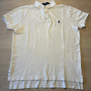 Vit pikétröja från Polo Ralph Lauren - Vit pikétröja från Polo Ralph Lauren i klassisk custom fit. Tröjan har korta ärmar, krage och knappar framtill. På bröstet finns den ikoniska broderade loggan i mörkblått. Materialet är mjuk bomull som andas och passar perfekt till en chill stil. Storlek M i väldigt fint skick.