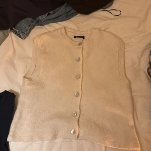 Beige stickad väst från Gina Tricot - Säljer en beige stickad väst från Gina Tricot i storlek M. Västen har rund hals, ribbade kanter och fem silvriga knappar framtill. Perfekt att styla över en skjorta eller t-shirt för en chill look.