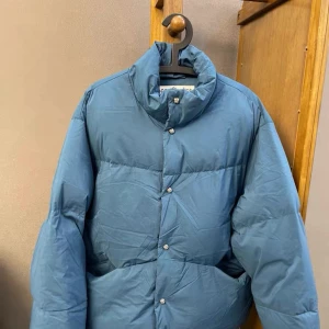 Blå pufferjacka från Acne Studios - Säljer en blå pufferjacka från Acne Studios med hög krage och snygga knappar framtill. Jackan har en clean look och är perfekt för kalla dagar. Materialet är mjukt och jackan har en oversized passform som ger en cool vibe.