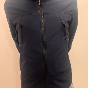 Mörkblå vindjacka från C.P. Company - Snygg mörkblå vindjacka från C.P. Company med huva och två snedställda fickor med dragkedja framtill. Jackan har en stilren design med hel dragkedja och diskret logga på huvan. Perfekt för dig som gillar streetwear och funktionella plagg.