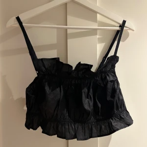 Svart volang linnetopp från H&M - Supersöt svart linnetopp från H&M med justerbara smala axelband och volangdetaljer både upptill och nedtill. Toppen är croppad och har en luftig, rynkad design som ger en cool och trendig vibe. Perfekt för att lyfta din outfit med lite extra edge.