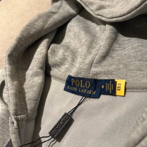 Grå hoodie från Polo Ralph Lauren - Snygg grå hoodie från Polo Ralph Lauren med dragkedja framtill, huva med snörning och klassisk broderad logga på bröstet. Tillverkad i mjuk bomullsmix, perfekt för chill dagar. Fickor framtill och ribbade muddar för extra komfort. Säljer för sparar pengar