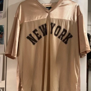 Champagnefärgad New York t-shirt från Cider - Snygg champagnefärgad t-shirt från Cider med 'NEW YORK' tryckt i brun-ish på framsidan. Oversized passform, korta ärmar och v-ringad hals. Övre delen är glansig medan nedre delen har meshstruktur. Perfekt för dig som gillar streetwear och trendig stil. Liknar Tate Mcrae och Billie Eillish style.  NYTT MED TAGS 