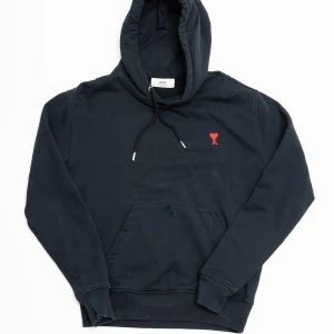 Svart hoodie från Ami med rött hjärta - Svart hoodie från Ami med klassisk huva och snörning. På bröstet finns ett broderat rött hjärta med bokstaven A. Tröjan har en stor magficka och ribbade muddar vid ärmslut och nederkant. Perfekt för dig som gillar stilrena detaljer. Några små hål på fickan.