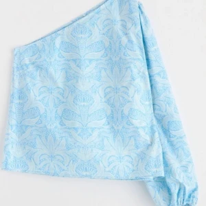 Blå oneshoulder topp med blommigt mönster - Säljer en ljusblå oneshoulder topp med ett blommigt mönster i vitt. Knappt använd så mycket fint skick 