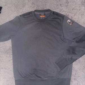 Mörkgrå fleece-tröja Parajumpers S - PRIS KAN DISKUTERAS VID SNABB AFFÄR. Mörkgrå långärmad fleece-tröja från Parajumpers i storlek S. Tröjan har rund hals, ribbade muddar och ett snyggt PJS-märke på ena ärmen. Diskreta röda sömmar vid axlarna ger en schysst detalj. Perfekt för dig som gillar stilrena och bekväma plagg.