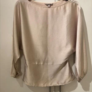 Champagnefärgad blus från H&M - Säljer en stilren champagnefärgad blus från H&M med lätt glansig yta och diskret struktur. Blusen har långärmade, lösa ärmar med resår vid ärmslut och en markerad midja baktill. Materialet är mjukt och består av polyester med lite stretch för extra komfort.