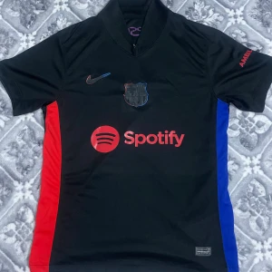 Barcelona kit 24-25 away kit  - Snygg FC Barcelona-tröja i svart med röda och blå detaljer på sidorna. Officiell Nike-design med Spotify som sponsor på framsidan och UNHCR-loggan på baksidan. Högkvalitativt material, perfekt passform och aldrig använd-helt ny.