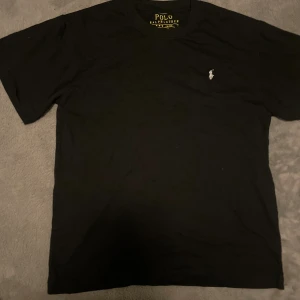 Svart t-shirt från Polo Ralph Lauren - Svart t-shirt från Polo Ralph Lauren med klassisk passform och korta ärmar. T-shirten har en liten vit broderad logga på bröstet och är tillverkad i mjuk bomull. Perfekt för dig som gillar stilrena och tidlösa plagg.