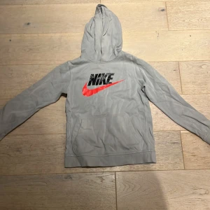 Grå Nike hoodie med röd swoosh - Snygg grå hoodie från Nike med stor svart logga och röd swoosh på bröstet. Klassisk känguruficka framtill och huva. Perfekt för chill eller träning. Standard fit och mjukt material.