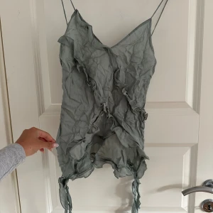 Grön omlottklänning från Zara - Supersnygg grön kortklänning från Zara med tunna axelband och v-ringad hals. Klänningen har volangdetaljer och omlott-effekt med knytband nedtill som ger en cool och trendig look. Perfekt för dig som gillar unika detaljer och en lätt, flowy känsla.