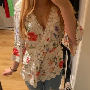 Blommig omlottblus med spetsdetaljer - Säljer en superfin blommig omlottblus i vit bas med röda, blå och gröna blommor. Blusen har spetsdetaljer längs v-ringningen och nederkanten, samt trekvartsärmar. Den har en luftig passform och är perfekt för vår och sommar. 