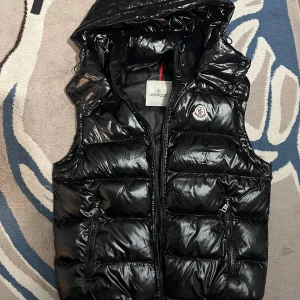 Moncler Väst - Storlek S Svart, glansig dunväst från Moncler med avtagbar huva och två dragkedjeförsedda fickor. Västen har quiltad design och Moncler-logga på bröstet. Perfekt för lager-på-lager och har en cool, urban vibe. Fodret har en unik serietidningsdetalj på insidan.