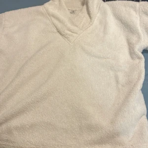 Vit fluffig fleecetröja med v-ringning - Supermjuk och mysig vit fleecetröja med v-ringad krage. Perfekt för kyliga dagar när du vill ha något riktigt bekvämt och varmt. Tröjan har lång ärm och är gjord i fluffigt fleecematerial som känns skönt mot huden.