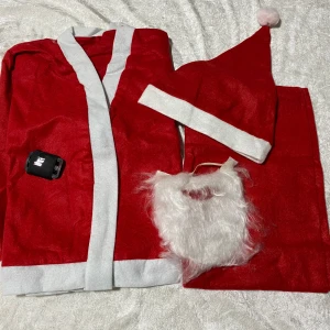 Barn Tomtedräkt med skägg och luva - Röd tomtedräkt med vita detaljer, inkluderar kappa, byxor, luva med vit boll och ett vitt skägg. Perfekt för julens alla upptåg och maskerader. klassisk tomtestil