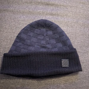 Mörkblå Louis Vuitton mössa i ull - Exklusiv mörkblå mössa från Louis Vuitton med diskret rutigt mönster och ribbad uppvikt kant. Mössan är tillverkad i mjuk ull och har en liten patch med Louis Vuitton-logga framtill. Perfekt accessoar för att lyfta din vinterstil.